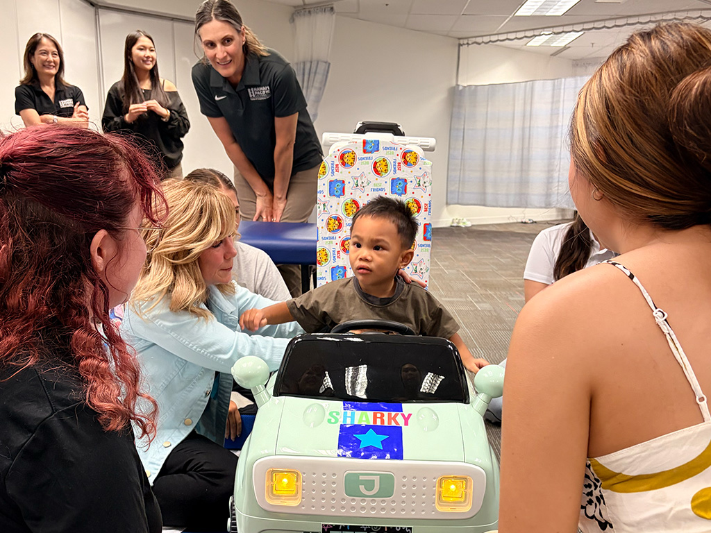 A child getting ready to drive his customized ride-on car. Standing behind the car is Jana Cason and kneeling with the child (left) is Patricia Coker-Bolt